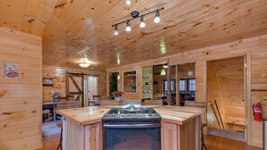 A wooden cabin interior with a central kitchen island, wooden walls, and a ceiling with exposed beams and lights.