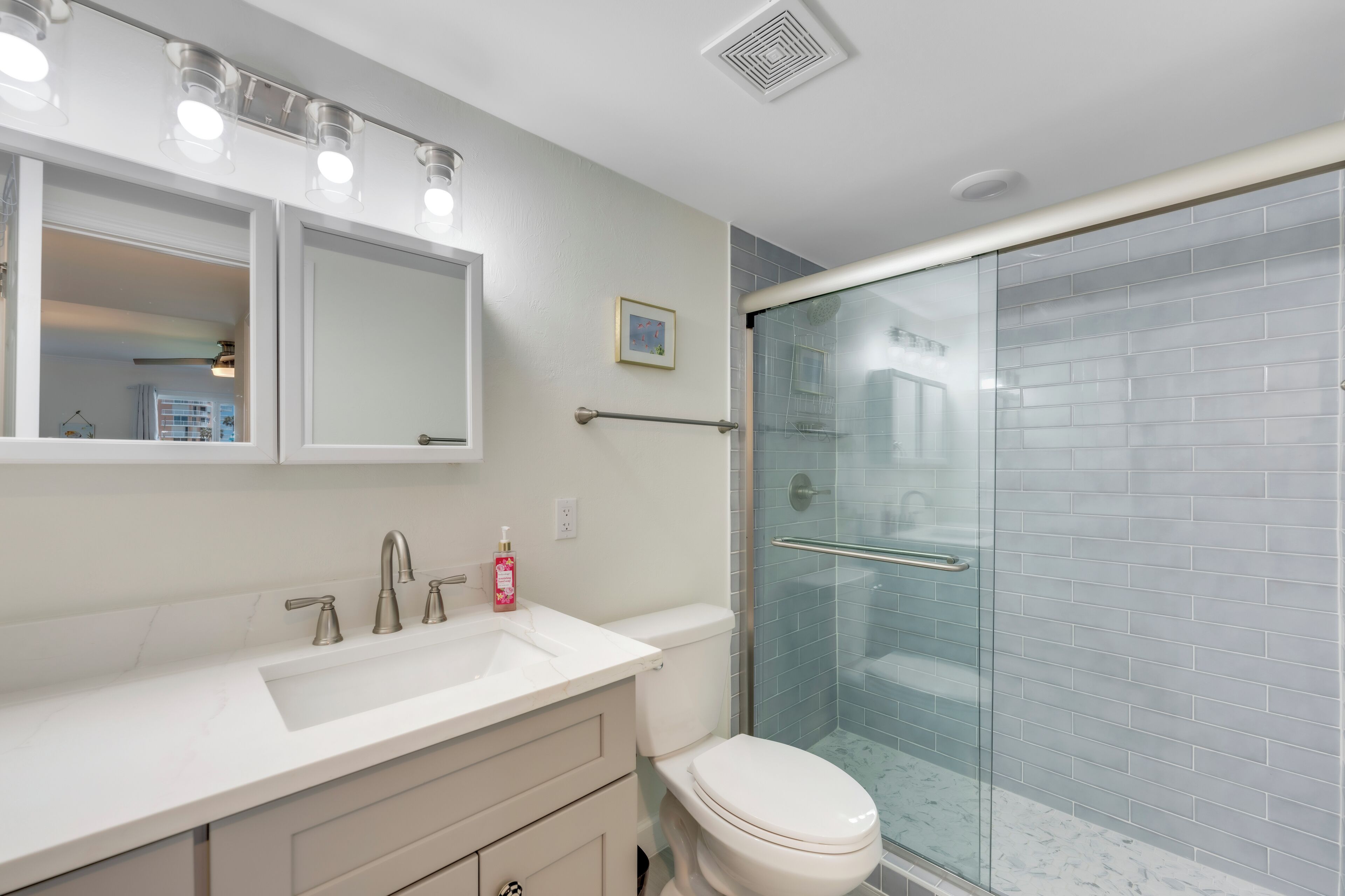 A modern bathroom with a white sink, vanity, and toilet. It includes a glass shower enclosure and a large mirror with lights above.