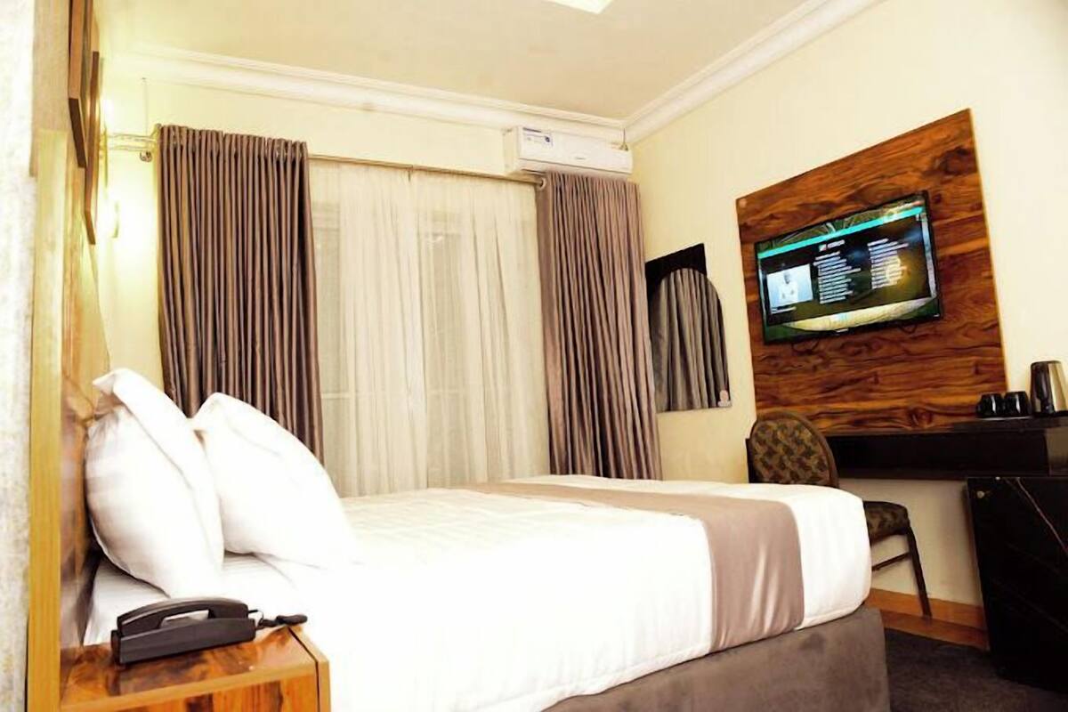 A hotel room with a bed, a desk, a chair, an air conditioning unit, and a television mounted on the wall.