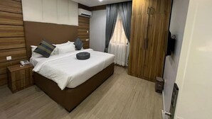 A modern bedroom with a large bed, wooden headboard, a bedside table, a nightstand, a wardrobe, and a ceiling light. - Clevero Hotel  (Abakaliki)