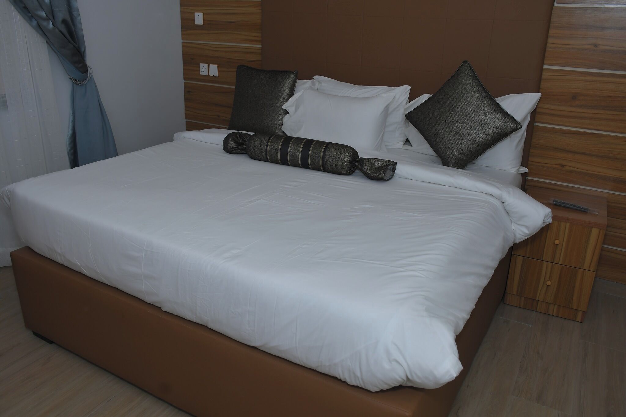 Een keurig opgemaakt bed met witte lakens, een bruin hoofdbord en een houten nachtkastje met lade.