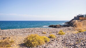 A rocky beach with a clear view of the ocean and a building with a red railing on the right. - Villa Luisa: Relax with ocean view and swimming pool (Los Abrigos)