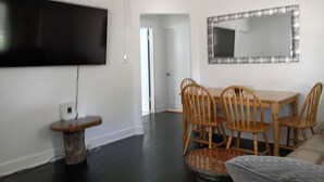 A modern living room with a flat-screen TV mounted on the wall, a dining table with chairs, and a large mirror above the table.