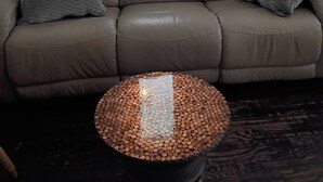 A beige sofa with two pillows, a round table with a copper-colored top, and a window with blinds.