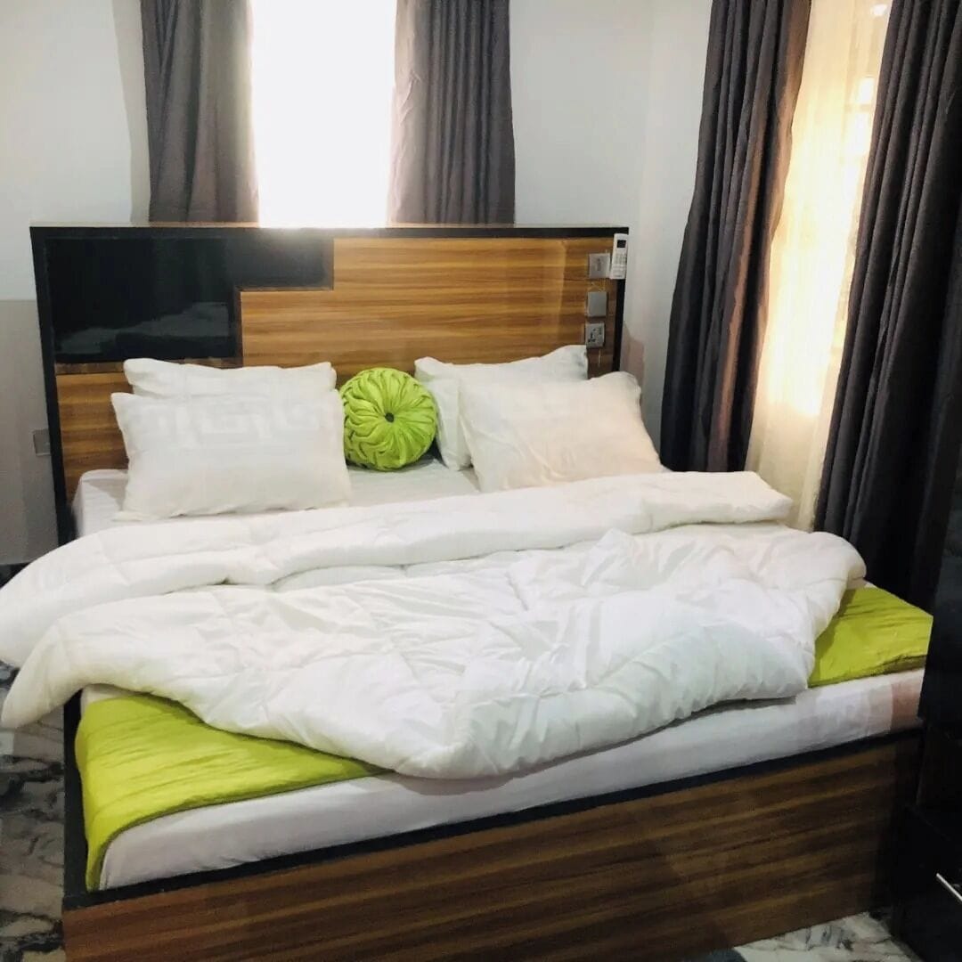 A neatly made bed with white bedding and a green pillow, a wooden headboard, and a marble floor.