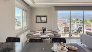 Un comedor con una mesa preparada para cuatro personas, un sofá y vistas a una ciudad y montañas a través de grandes ventanales.