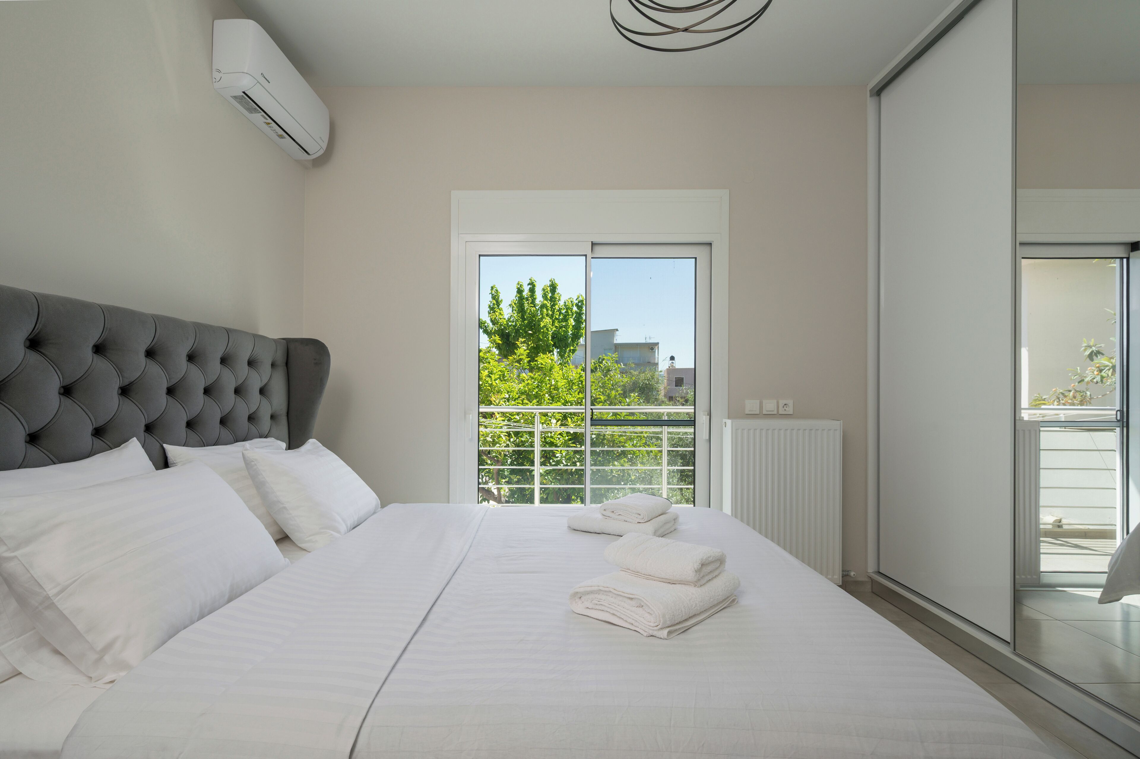 A neatly made bed with white linens and towels, a tufted headboard, and a view of greenery through a window.