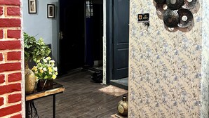Ein Flur mit Backsteinwand, Holzboden und einer Tür mit Blumenmuster. Ein Tisch mit Blumen und einer Ziervase steht dort.