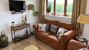 A cozy living room with a brown leather sofa, a wooden console table, a television mounted on the wall, and a window with curtains.