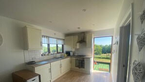 A modern kitchen with white cabinets, a stove, and a sink. There is a brown sofa in the living area and a view of a garden through the open door.