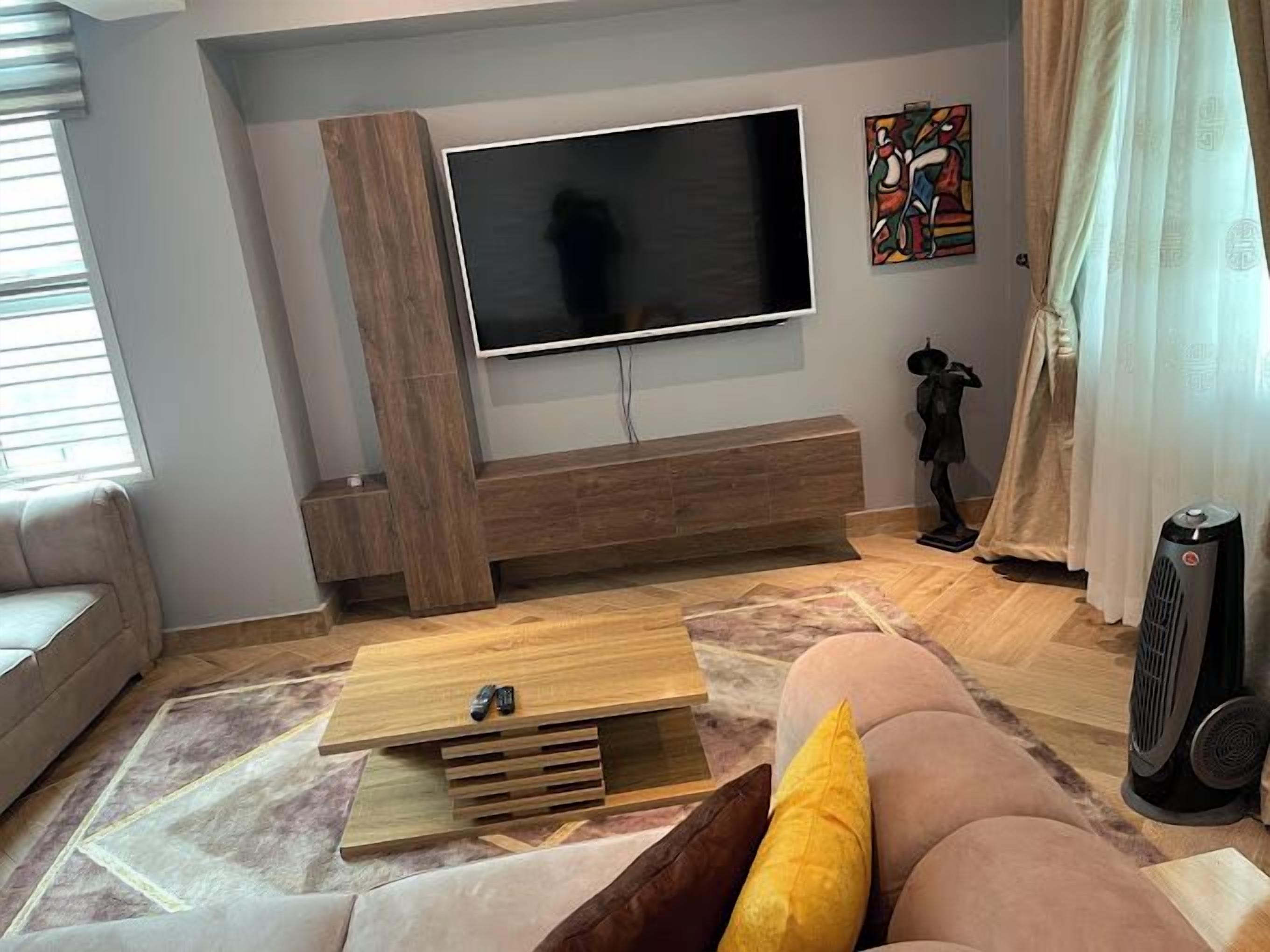 En moderne stue med et fladskærms-tv monteret på en trævægsenhed, en beige sofa, et træsofabord og en tæppe med et mønster.