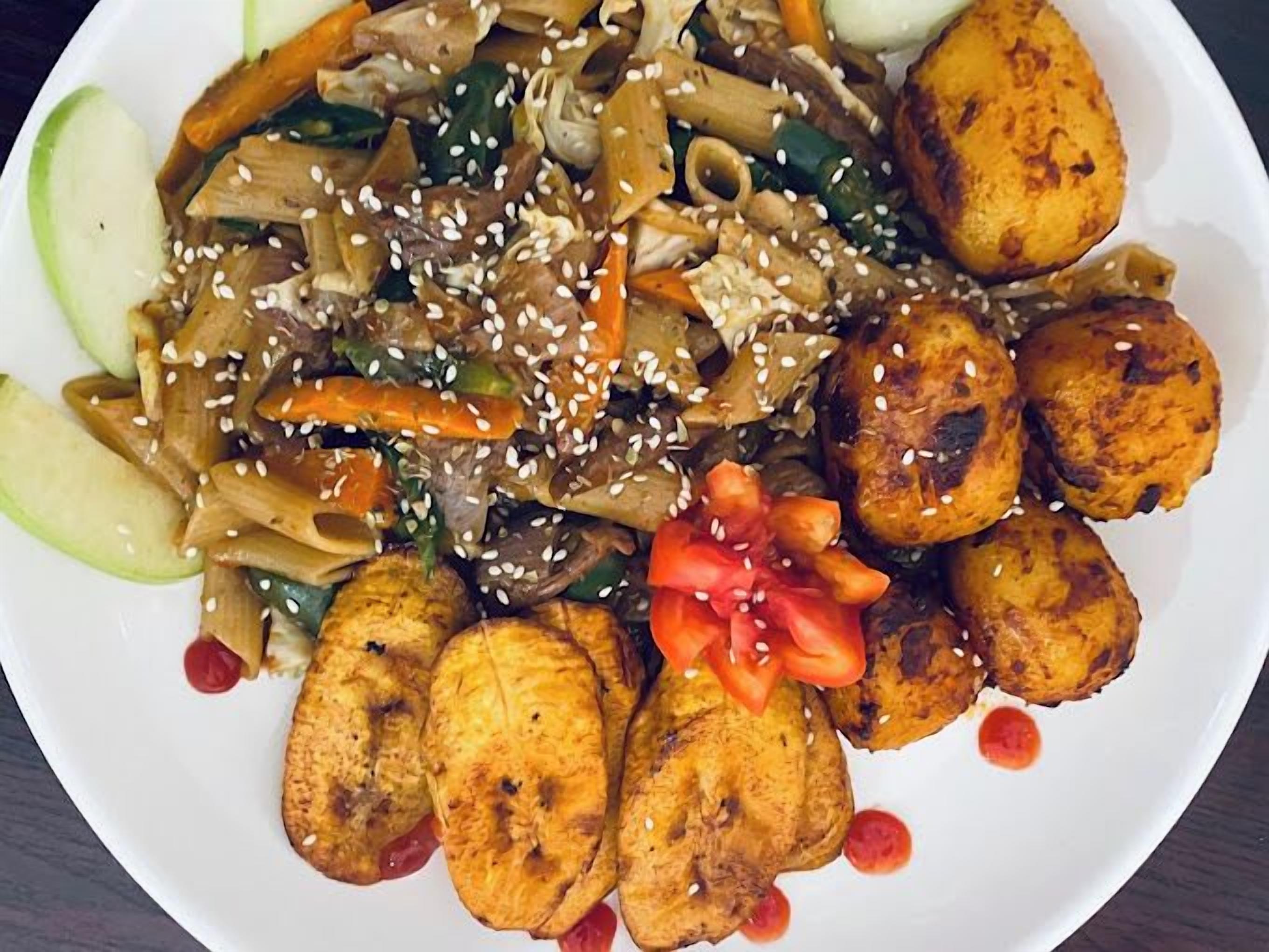 Eine Portion gebratener Nudeln mit Gemüse und Tofu, garniert mit Sesamsamen, dazu gebratene Kochbananen und Gurkenscheiben.