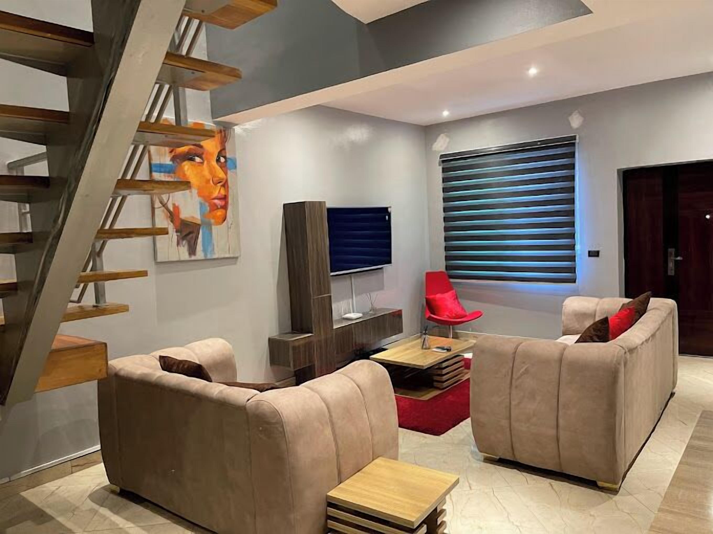 A modern living room with a staircase, a flat-screen TV, a red chair, and a sofa set.