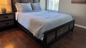A bedroom with a wooden bed frame, a nightstand, a lamp, and a window with blinds.