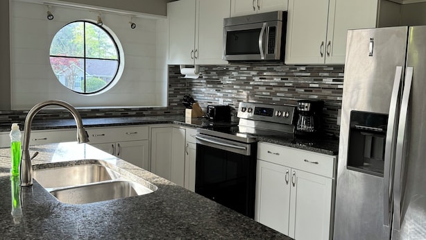 A modern kitchen with white cabinets, stainless steel appliances, a granite countertop, and a window with a view of trees.