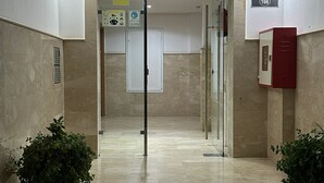 Un couloir doté d’un sol en marbre, de portes en verre et de plantes en pot. Une enseigne est placée au-dessus de la porte et une boîte électrique rouge est fixée au mur.