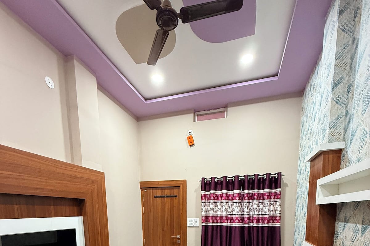 Une chambre à coucher comprenant un lit, un téléviseur, un ventilateur de plafond, une porte et un rideau à motifs.