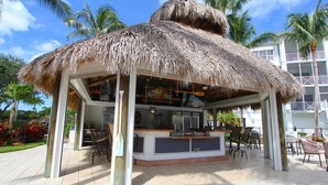 A thatched-roof outdoor bar area with seating, a counter, and a view of a multi-story building in the background. - Tropical Poolside Retreat! (Bonita Springs)
