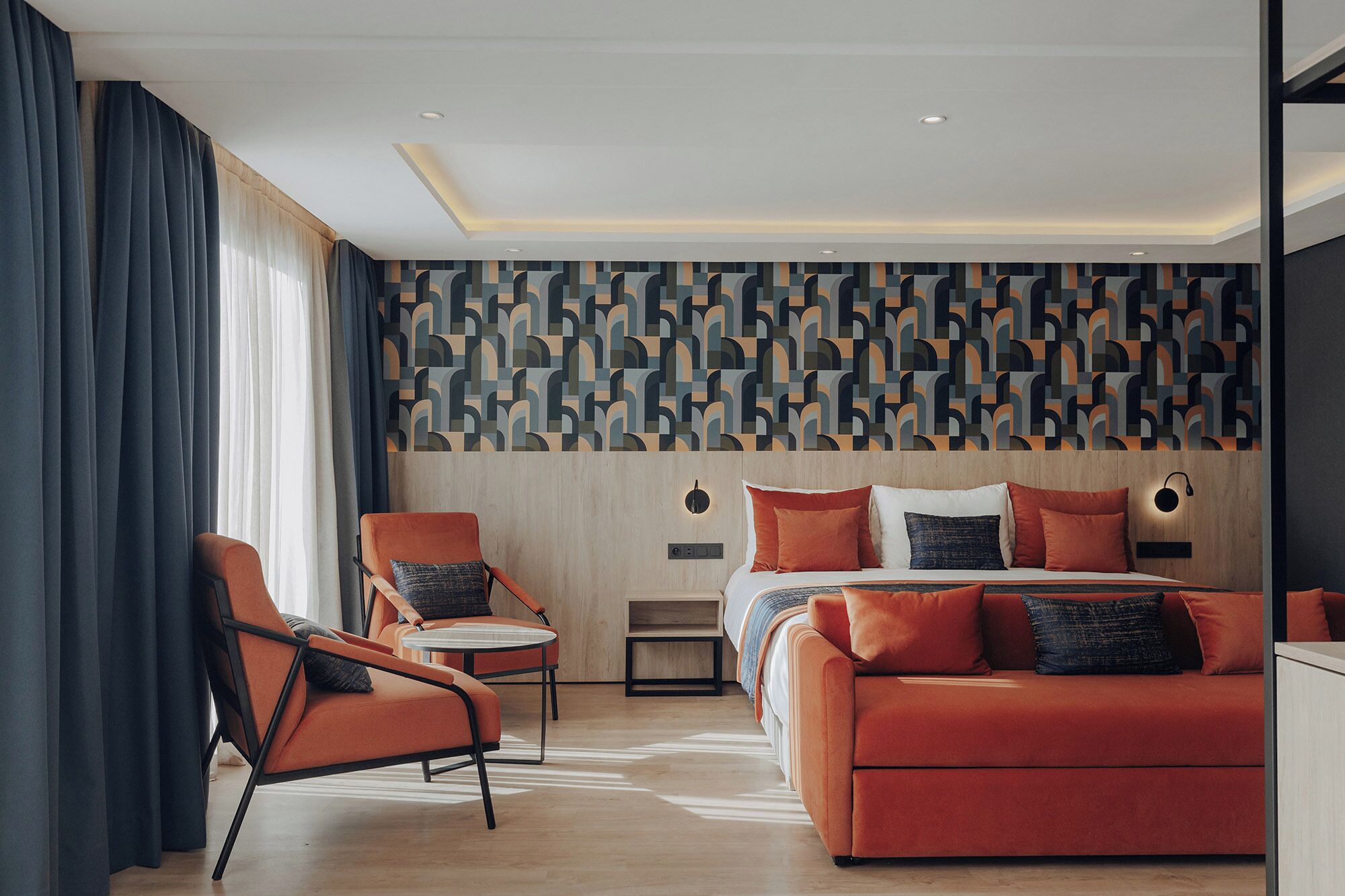 A modern hotel room with a geometric patterned wall, a bed with multiple pillows, an orange sofa, and a small round table.