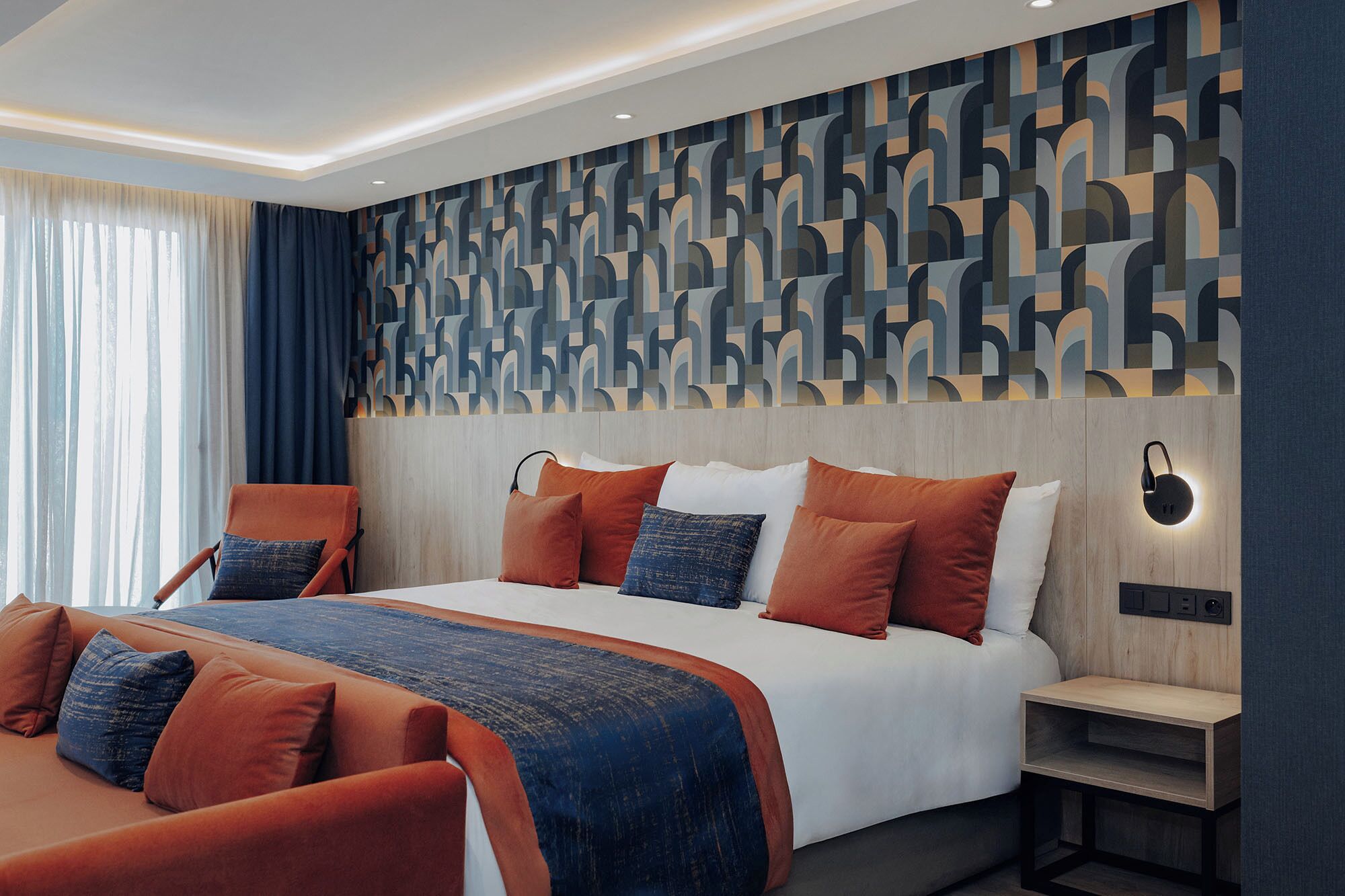 A modern hotel room with a large bed, two armchairs, a nightstand, and a patterned wall.