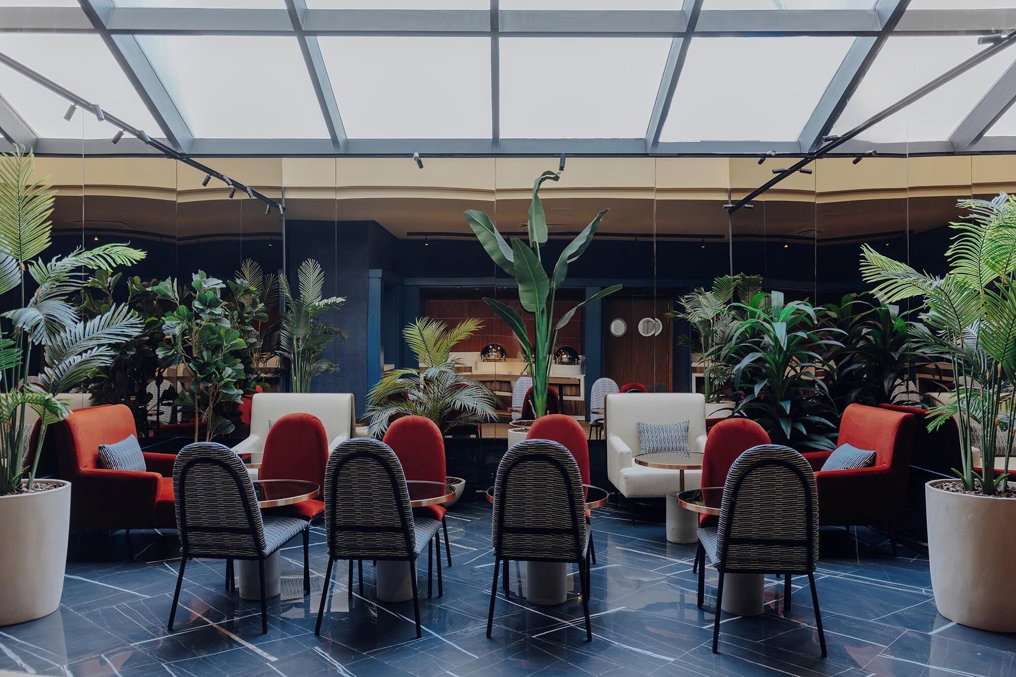 A spacious indoor area with a high ceiling and skylight, featuring multiple seating arrangements and abundant indoor plants.