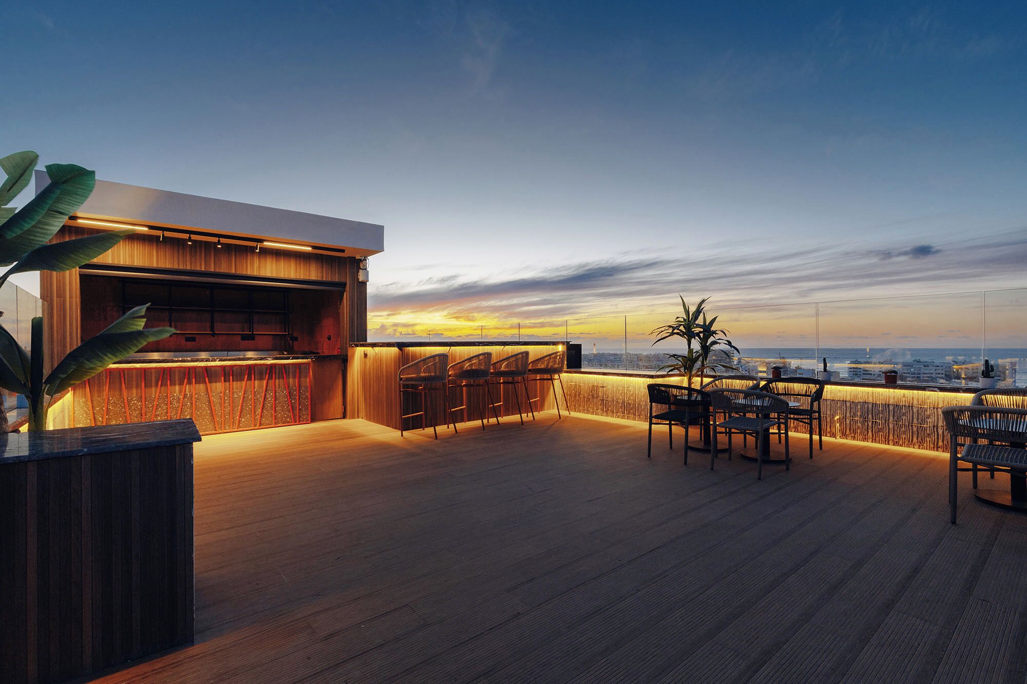 A rooftop bar with a wooden floor, bar counter, and seating area with chairs.