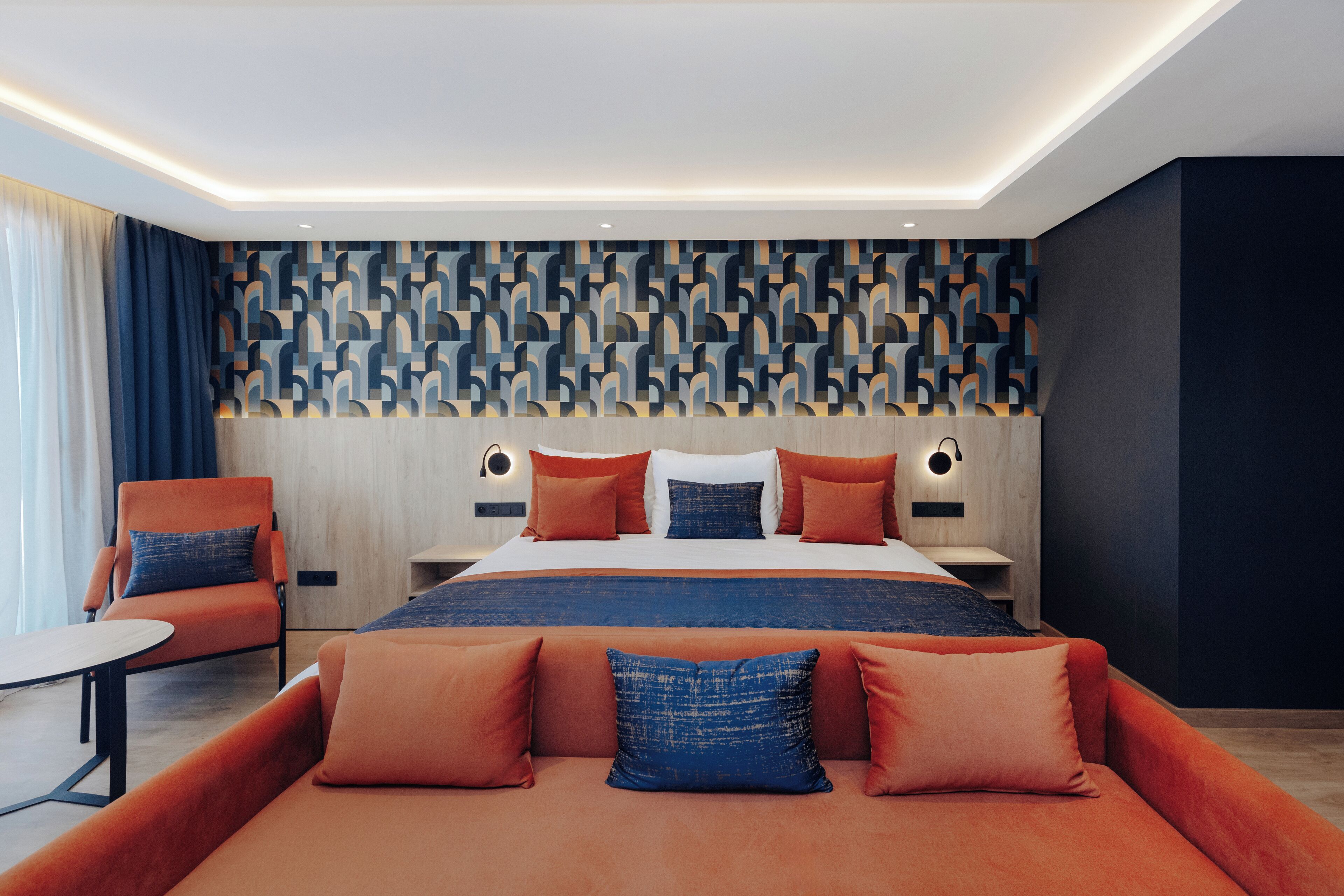 Una habitación de hotel moderna con una cama grande, ropa de cama naranja y azul, un cabecero de madera y un muro con estampado geométrico.