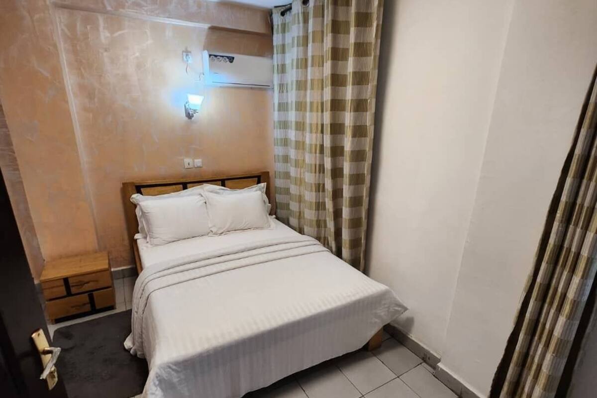 A bedroom with a bed, a nightstand, a wall-mounted air conditioner, and a window with curtains.