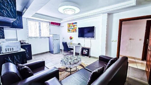A modern living room with a sofa set, a coffee table, and a TV. There is a kitchen area with a microwave and a refrigerator. - RESIDENCE FAMILIA PHOENIX (Douala)