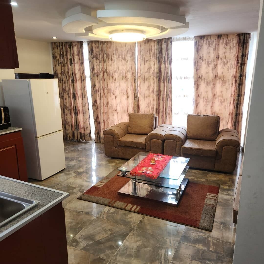 A modern living room with a glass coffee table, two sofas, a kitchen area with a refrigerator, and a large window with curtains.