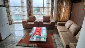 A living room with a sofa set, a glass coffee table, and a view of the city through large windows. - RESIDENCE FAMILIA PHOENIX (Douala)