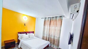 A bedroom with a bed, a nightstand, a wall-mounted air conditioner, and a window with curtains. - RESIDENCE FAMILIA PHOENIX (Douala)
