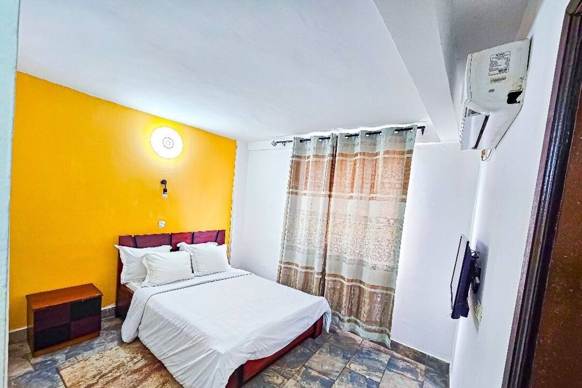 A bedroom with a bed, a nightstand, a wall-mounted air conditioner, and a window with curtains.