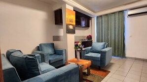 A living room with two blue sofas, a small table with a vase of flowers, and a kitchen area in the background. - RESIDENCE FAMILIA PHOENIX (Douala)