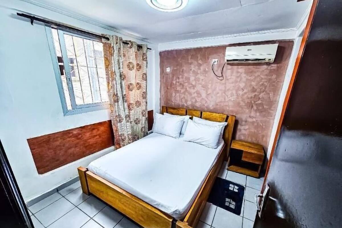 A bedroom with a wooden bed, a window with curtains, an air conditioning unit, and a tiled floor.