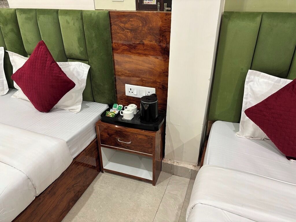 A hotel room with two beds, each with a green upholstered headboard, white bedding, and red pillows. A wooden nightstand with a coffee maker and a small table with a green cup and a white mug is between the beds.