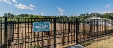 A fenced dog park with a sign, a paved walkway, and a building in the background.