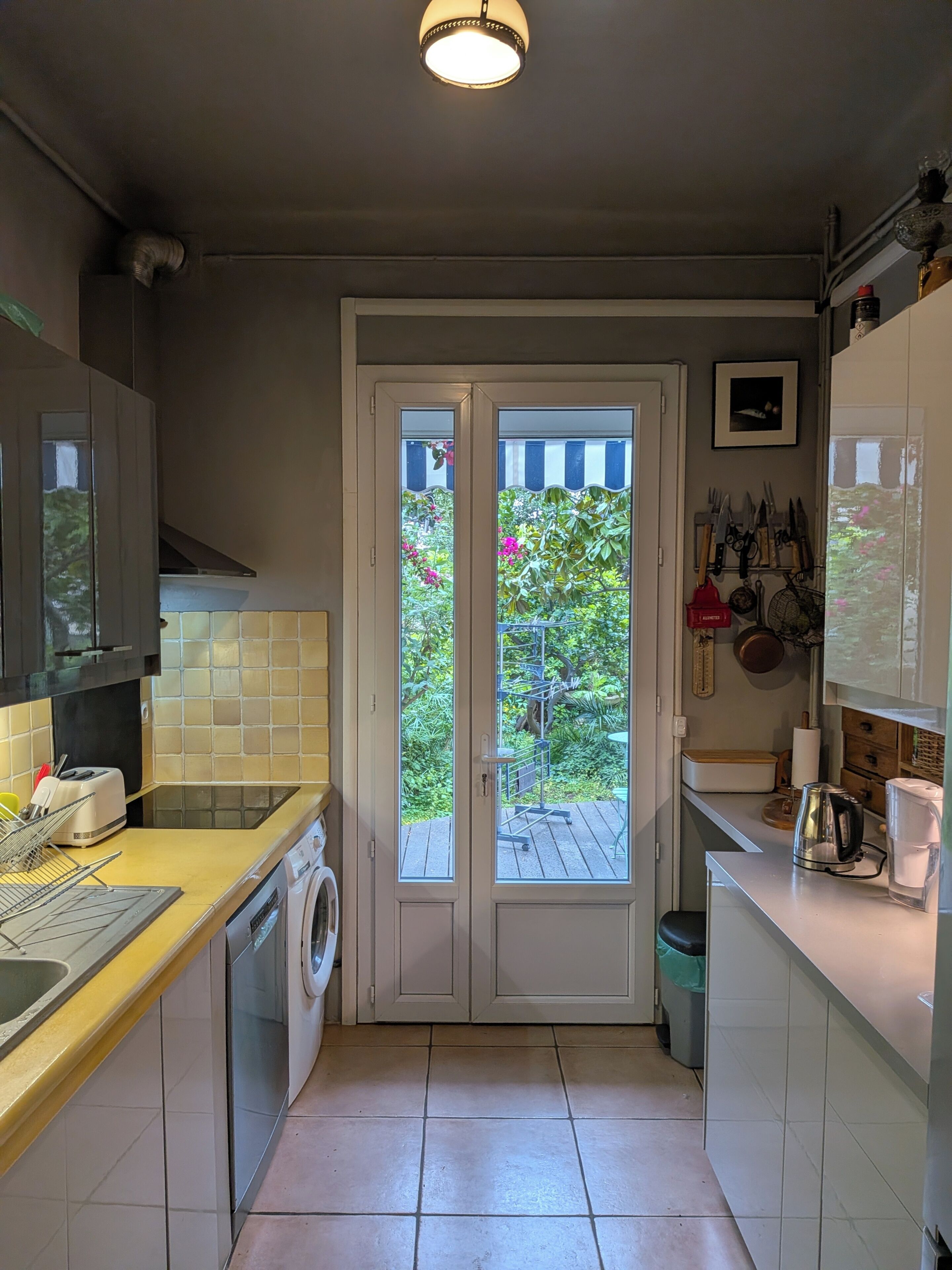 Une cuisine compacte équipée d’un lave-linge, d’un grille-pain et d’un évier. Une porte donne accès à un balcon agrémenté de plantes et offrant une vue sur la verdure.