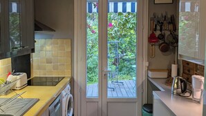 A compact kitchen with a washing machine, a toaster, and a sink. There is a door leading to a balcony with plants and a view of greenery.