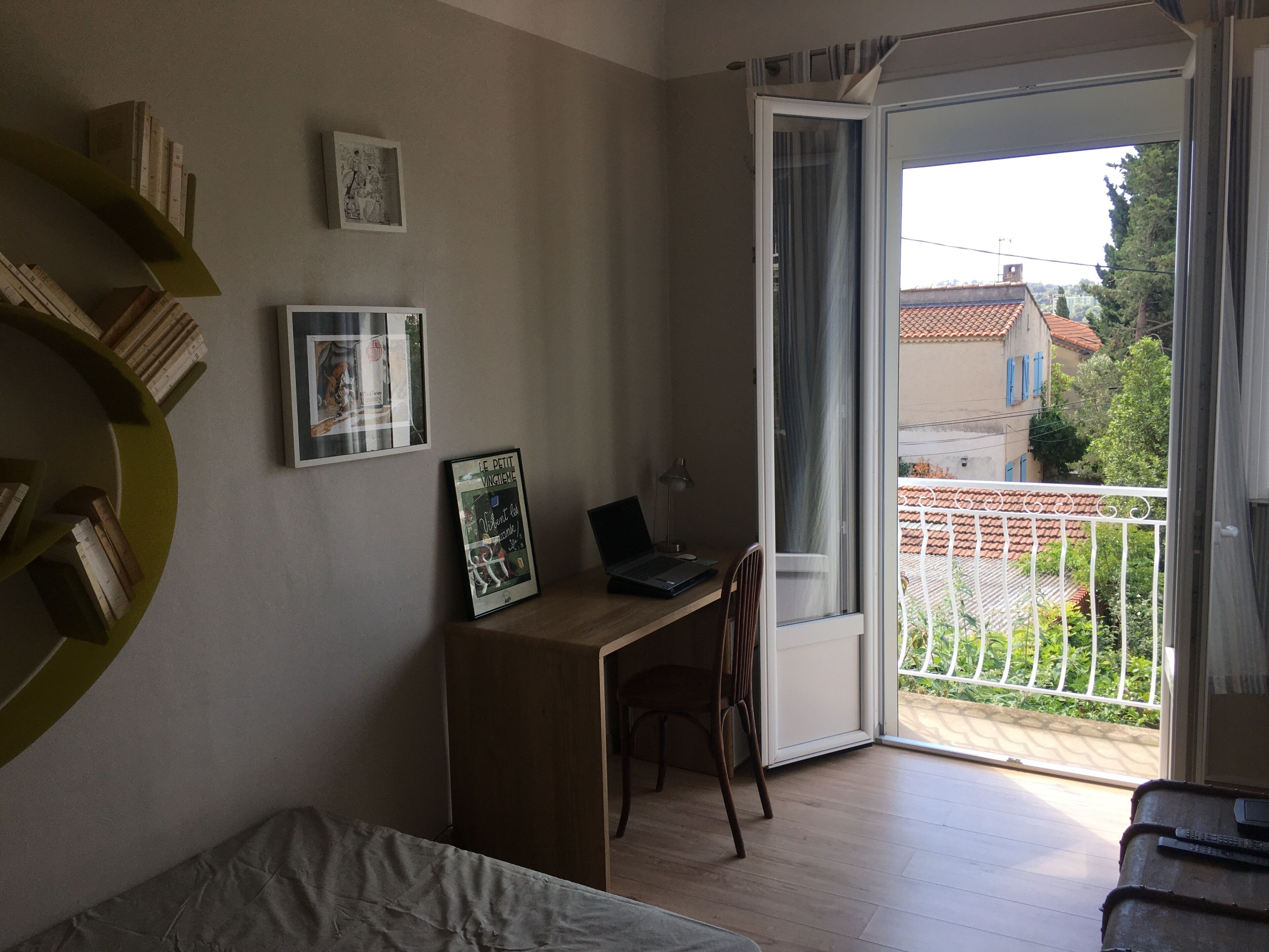 Una habitación con escritorio de madera, una computadora portátil, una silla, una cama y un balcón con vista a edificios y vegetación.