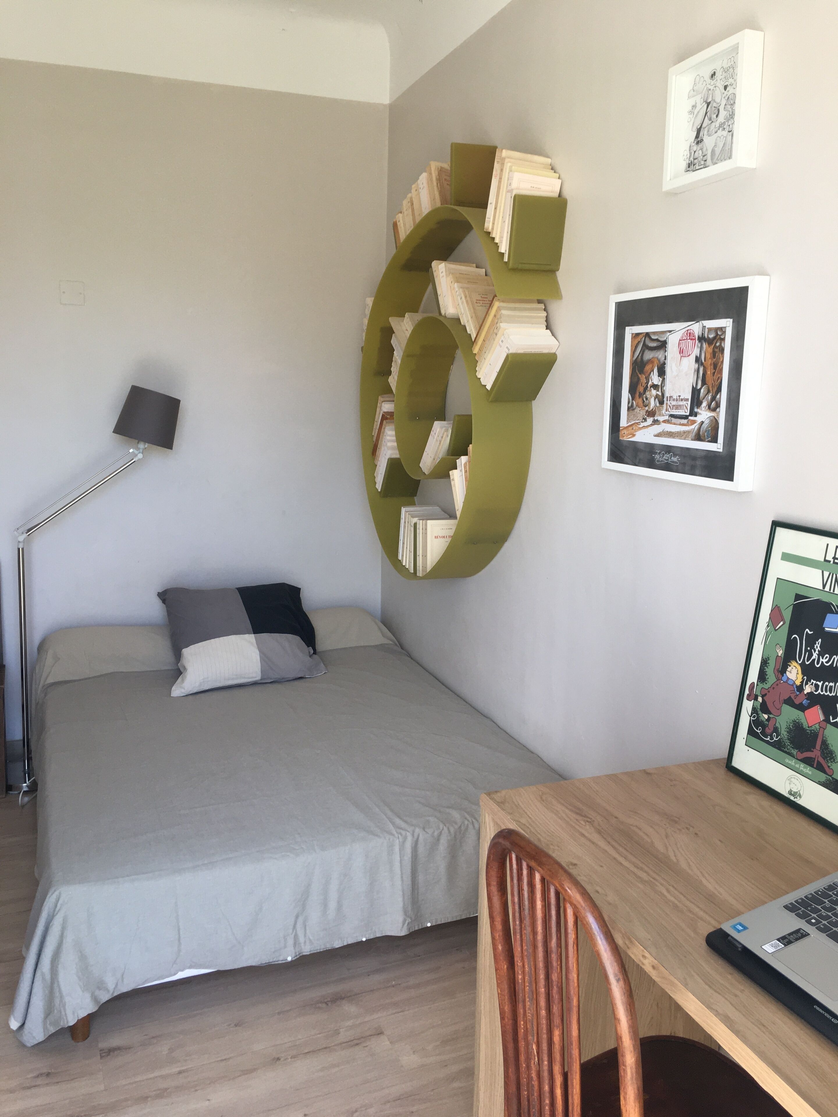 A bedroom with a bed, a desk with a laptop, a chair, a floor lamp, and a bookshelf.