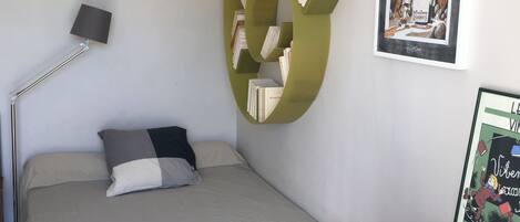 A bedroom with a bed, a desk with a laptop, a chair, a floor lamp, and a bookshelf.