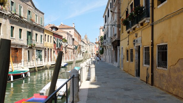 A narrow canal with boats docked on the left, flanked by historic buildings with balconies and windows.