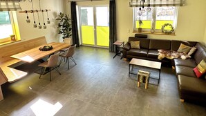 Moderni olohuone, jossa on puinen ruokapöytä, sohva ja sohvapöytä. Ikkunalaudalla on kasvi ja pieni pöytä, jossa on lamppu.