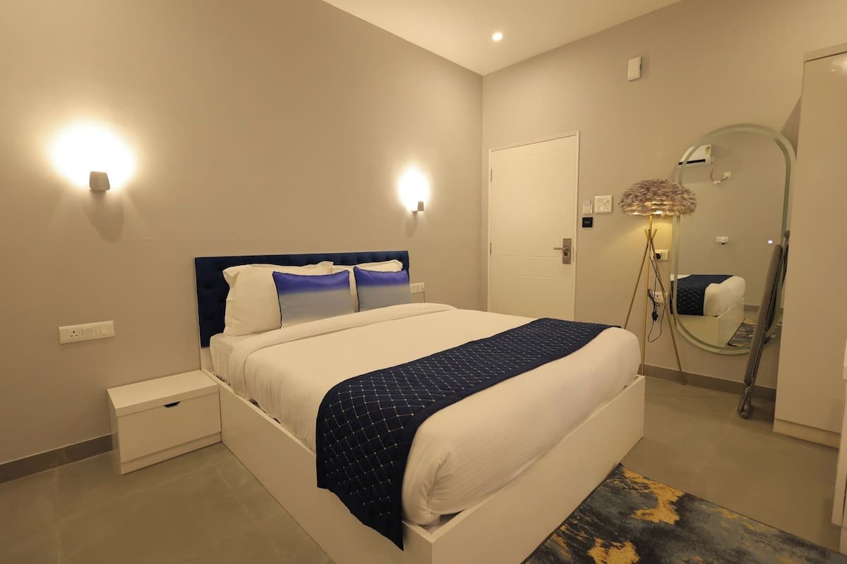 A hotel room with a bed, bedside table, wall-mounted lights, a floor lamp, a mirror, and a door.