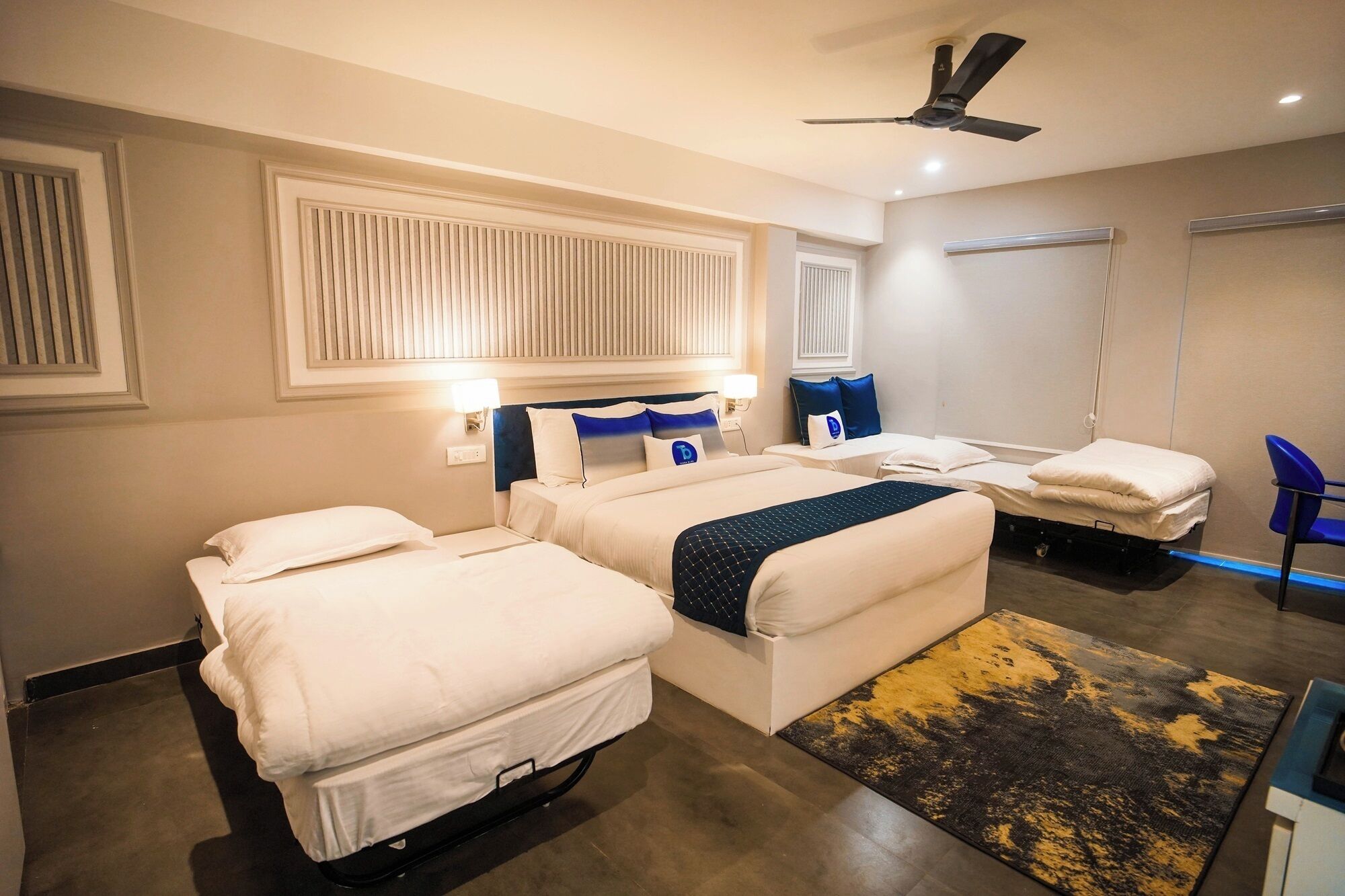Una habitación de hotel con dos camas, un ventilador de techo, una mesita de noche con lámpara y una silla azul.
