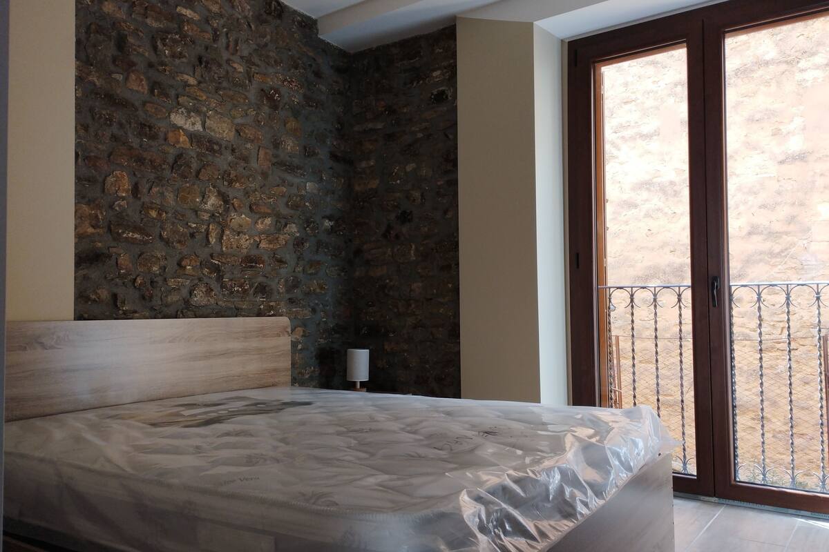 A bedroom with a stone wall, a bed with a wooden headboard, a nightstand, and a view of a balcony.