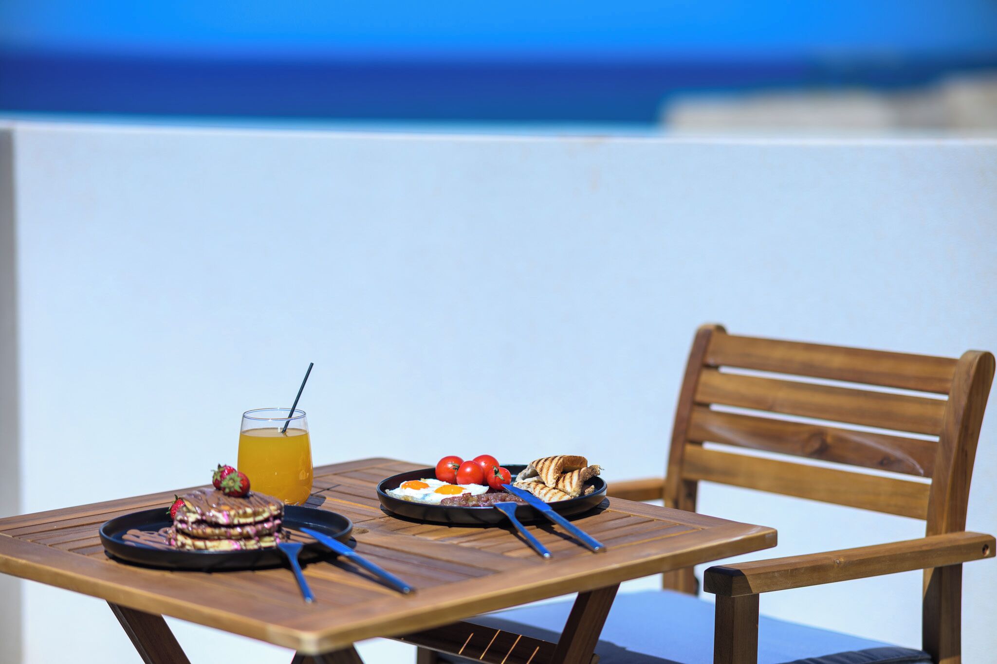 A wooden table with breakfast items including a plate of eggs, tomatoes, and croissants, and a glass of orange juice.