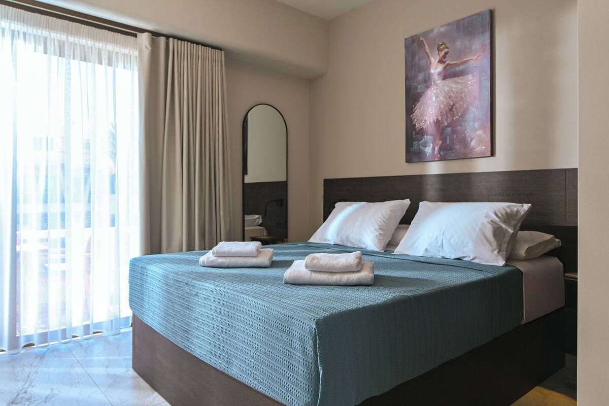 Habitación de hotel con una cama grande, un cuadro de una bailarina, un espejo y vistas de un edificio a través de la ventana.