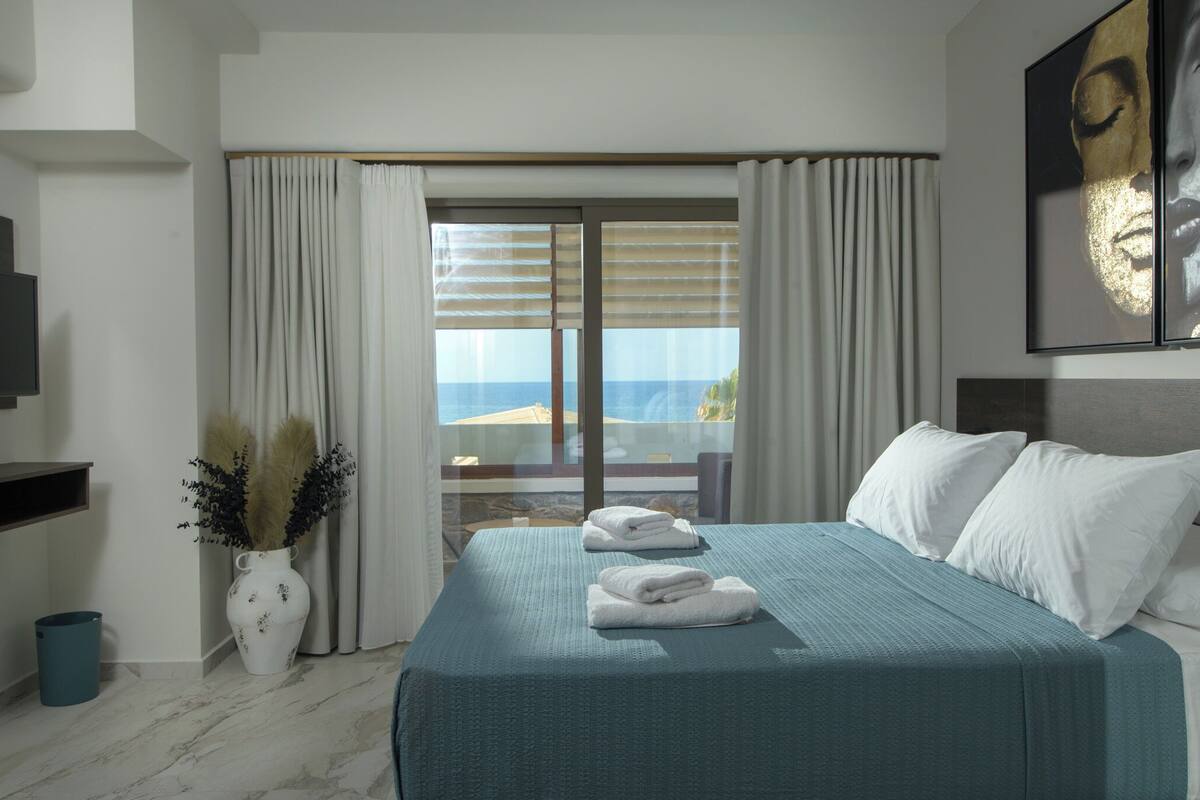 Habitación de hotel con cama, televisor, un jarrón con plumas de mispo y vistas al mar a través de la ventana.